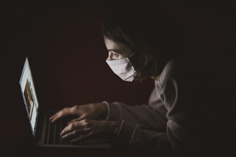 Osoba siedzi przy komputerze z ubraną maseczką na ustach